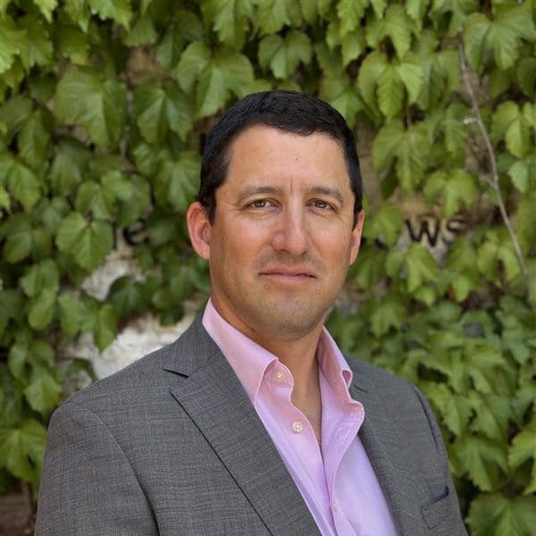 A man in a gray suit and pink shirt stands in front of a wall covered with green ivy leaves, looking at the camera with a neutral expression.